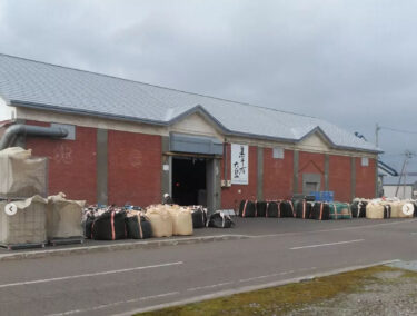 कुरोसेंगोकू सोयाबीन की कटाई कंटेनरों और थैलियों में भरकर होकुर्यु कस्बे में लाई जाती है और कारखाने की दीवारों पर कतार में लगा दी जाती है। [कुरोसेंगोकू व्यापार सहकारी संघ]